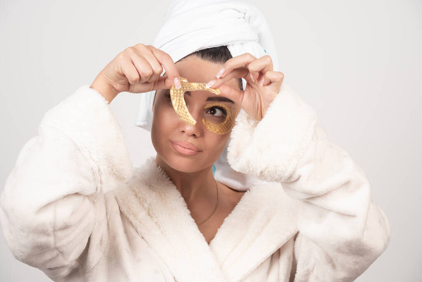 Genç esmer kadın bornoz giyiyor kozmetik göz bantlarını çıkarıyor. Yüksek kalite fotoğraf - Fotoğraf, Görsel