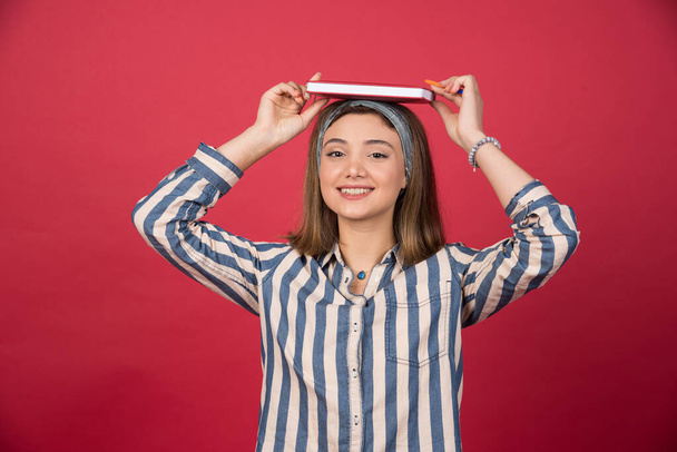 Chica joven pone libro en la parte superior de su cabeza sobre fondo rojo. Foto de alta calidad - Foto, Imagen