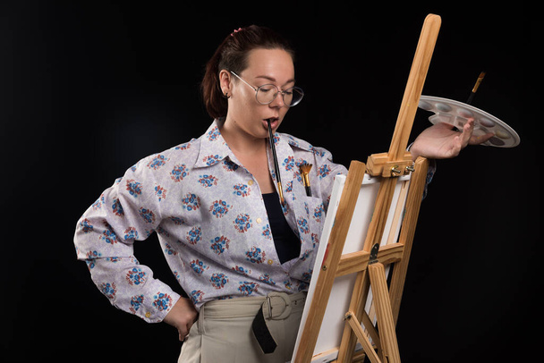 Woman artist holding brush and looking at canvas on black background  - Φωτογραφία, εικόνα