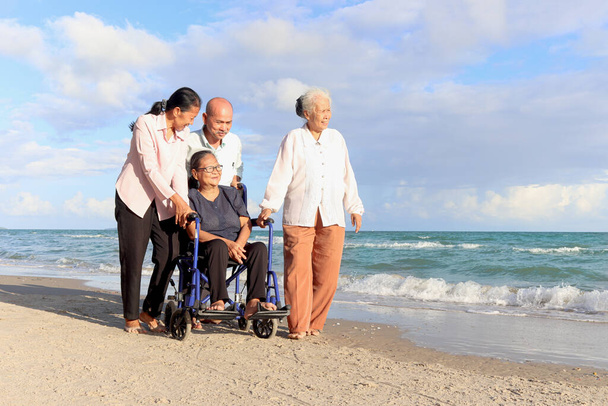 Happy disabled senior woman in wheelchair spending time with her friends on tropical sand beach. Group of four cheerful Asian elderly old friends travel outdoor together. - Photo, Image