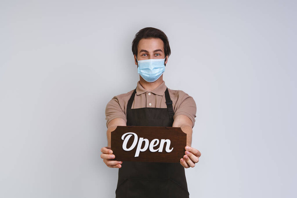 Handsome young man in apron wearing protective face mask and holding open sign while standing against gray background - Foto, immagini