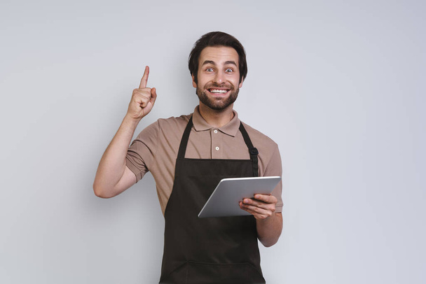 Cheerful young man in apron showing his digital tablet and gesturing while standing against gray background - Fotografie, Obrázek