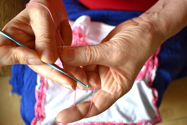 Oudere vrouw maakt haar borduurwerk en breien met een roze en witte draad en een paar naalden - Foto, afbeelding
