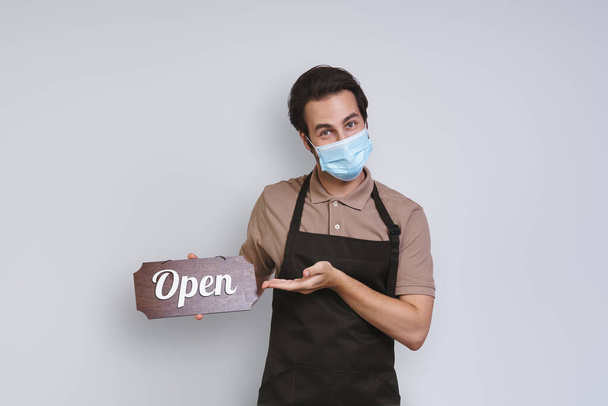 Handsome young man in apron wearing protective face mask and holding open sign while standing against gray background - Foto, Imagem