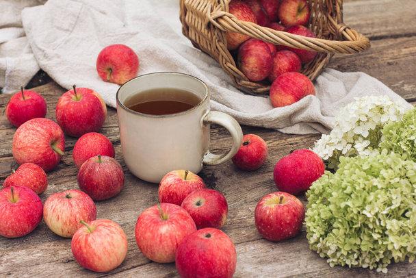 Autumn still life with a cup of tea, fresh red apples and hydrangea flowers on a textured wooden background. Autumn day and image, autumn photography, food decor, autumn light - Foto, Imagem
