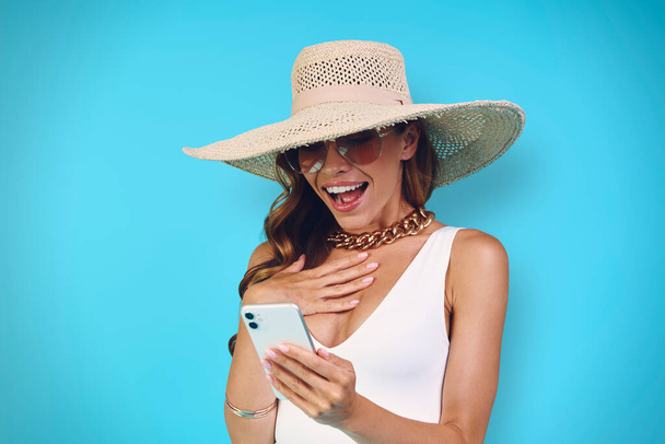 Meglepett fiatal nő elegáns kalap nézi a mobiltelefon, miközben áll szemben a kék háttér - Fotó, kép