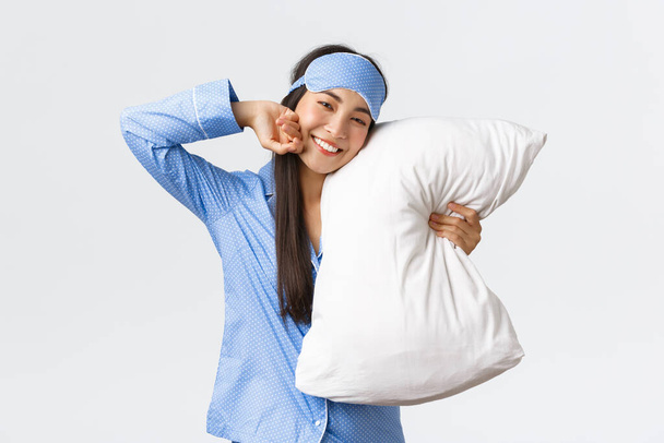 Pleased beautiful asian girl in blue pyjamas and sleeping mask, lying in bed and hugging pillow, smiling from satisfaction as stretching and feeling good after night sleep, white background - Photo, Image