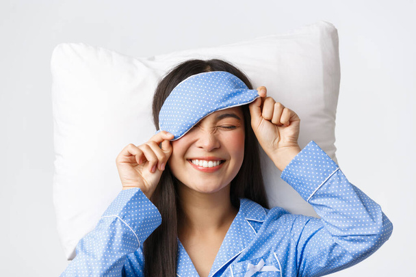 Zadowolony szczęśliwy uśmiechnięty koreański dziewczyna w niebieskiej piżamie i masce snu, miał wielki dzień idzie do łóżka w idealnym nastroju, przygotowując się do snu, leżąc na poduszce optymistycznie nad białym tle - Zdjęcie, obraz