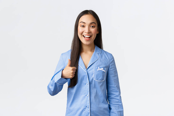 Upbeat happy asian pretty girl in blue pajamas smiling pleased and showing thumbs-up in approval or like, recommend product, great quality, showing well done or good job, white background - Фото, изображение