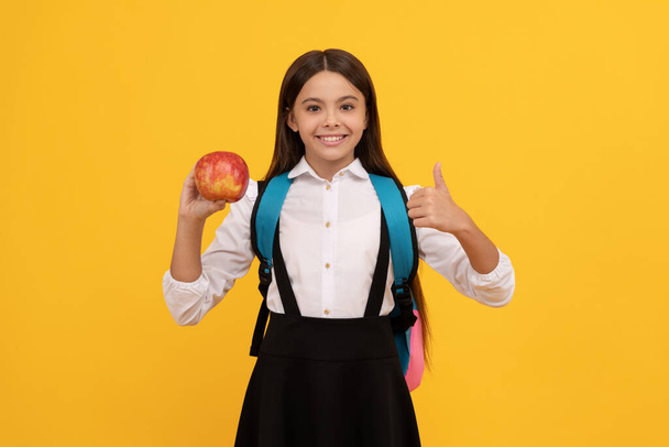 Счастливые девочки отдают большие пальцы, держа яблоко и школьный портфель на желтом фоне, образование - Фото, изображение