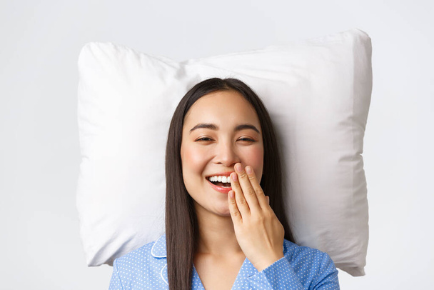 Χαμογελώντας χαρούμενη όμορφη Ασιάτισσα ξαπλωμένη στο κρεβάτι πάνω σε μαξιλάρι με μπλε πιτζάμες, ξυπνώντας ανοιχτά μάτια και χασμουρημένη, πρωινή ρουτίνα της νεαρής γυναίκας. Χαριτωμένο θηλυκό σε jammies διαμονή κρεβάτι, λευκό φόντο - Φωτογραφία, εικόνα