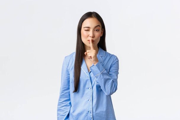 Vonzó ázsiai lány kék pizsamában rejteget valamit, kacsint ravasz és shushing, azt mondják, shhh mint kéri maradj csendben, csendben gesztus, miközben áll a fehér háttér felett ottalvós party - Fotó, kép