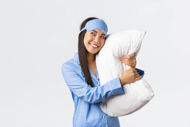 Мрійлива красива азіатська дівчина в блакитній піжамі і спальна маска обіймає подушку і усміхається задоволена, як виглядає верхній лівий кут зображення щось в ліжку, фотографування, використовуючи уяву
 - Фото, зображення