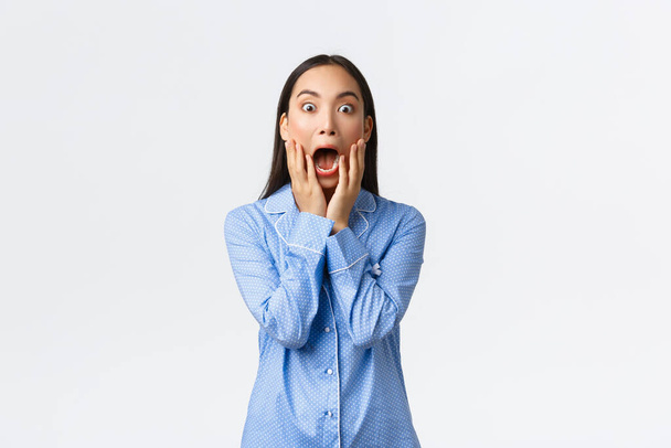 Surprised and shocked asian girl in blue pajamas screaming in awe and amazement, holding hands over opened mouth and stare at camera impressed, standing white background - Foto, Bild