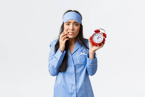 Anxious cute asian girl showing alarm and panic, biting finger concerned, looking nervous, dont know what do for being late, overslept to work in morning, wearing pajamas and sleeping mask - Photo, Image