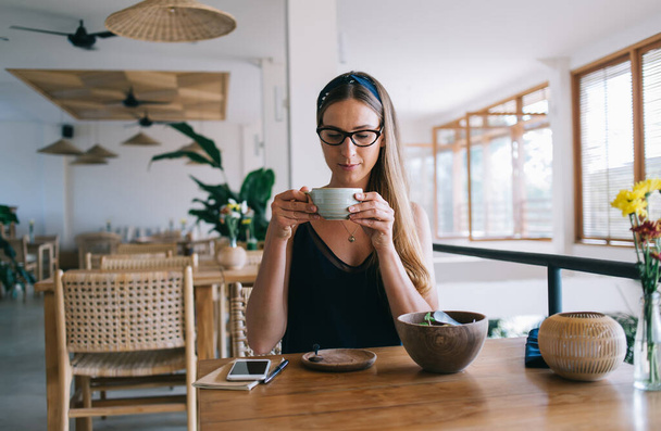 Pensive caucasian female in casual wear sitting at wooden table in cafeteria holding cup with beverage on breakfast, attractive woman enjoying caffeine beverage having lunch in cozy cafeteria - Foto, imagen