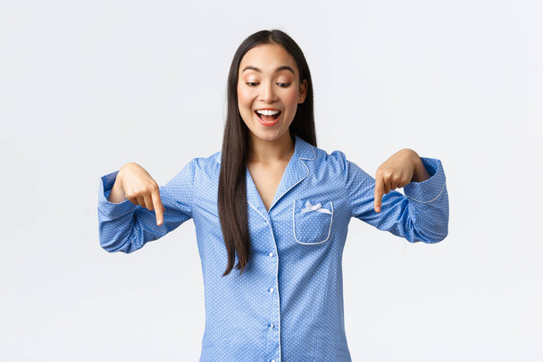 Entusiasta felice ragazza asiatica reagire alle notizie meravigliose, in piedi in pigiama blu e puntando il dito verso il basso felice, sorridente interessato e allegro, vedendo grande sconto promozionale, sfondo bianco - Foto, immagini