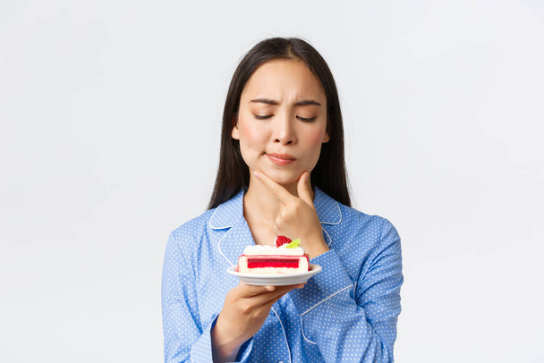 Close-up tiro de ponderando sério-olhando menina asiática no pijama olhando perplexo no bolo como pensar o quanto de calorias na sobremesa, querendo comê-lo à noite, de pé fundo branco
 - Foto, Imagem