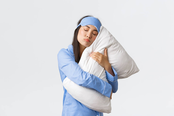 Silly and cute asian girl in blue sleeping mask and pajamas, hugging pillow tight as unwilling wake-up, dont want get up from bed in morning, heavy sleeper posing over white background - Foto, Imagen