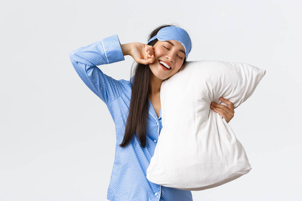 Smiling pleased cute asian girl in blue pyjama and sleeping mask, hugging pillow and stretching hands delighted as finally going bed, want sleep or waking up in morning, white background - Fotó, kép