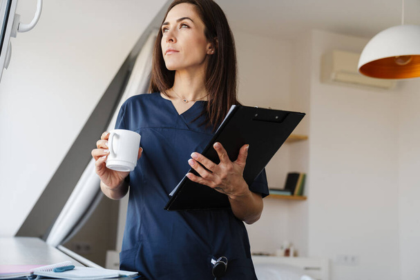 A pensive woman in a medical uniform standing near the window, holding a tablet with documents and a cup in her hands and looking aside in the room - Foto, Imagem