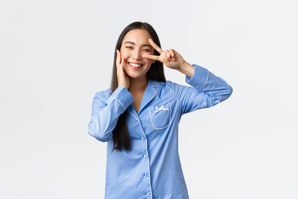 Γοητευτική ανόητη Ασιάτισσα με μπλε πιτζάμες που δείχνει το σημάδι ειρήνης και συγκινητικό μάγουλο kawaii, κοκκίνισμα και χαμόγελο, ικανοποιημένη με καθαρό τέλειο δέρμα, χρησιμοποιήστε προϊόντα περιποίησης δέρματος, λευκό φόντο - Φωτογραφία, εικόνα