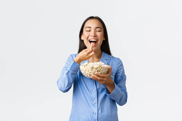 Home leisure, sleepover and slumber party concept. Smiling delighted asian girl enjoying eating favorite popcorn, wearing pajama on weekend, watching favorite movie, white background - Photo, image
