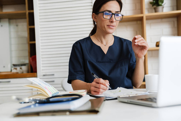 A woman in a blue medical suit and glasses sitting at a table in front of a laptop and looking into it in a bright room - 写真・画像