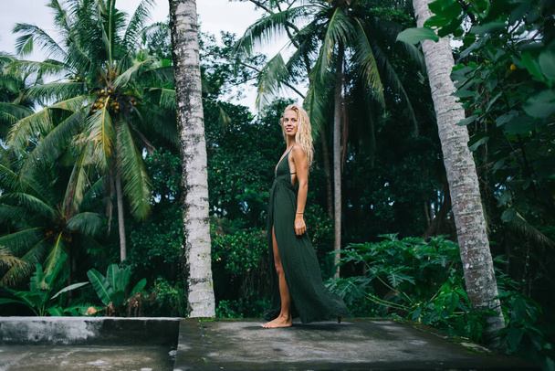 Beautiful tanned barefooted young lady with blonde fluffy fluffyhair wearing long flowing green dress and bracelet standing on background of tropical trees - 写真・画像