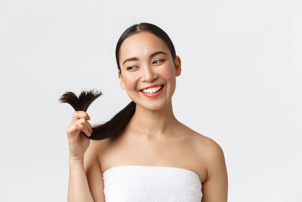 Beauté, produits de perte de cheveux, shampooing et concept de soins capillaires. Superbe femme asiatique heureuse dans une serviette de bain montrant les extrémités des cheveux et souriant satisfait, guéri pointes fourchues et regardant satisfait - Photo, image