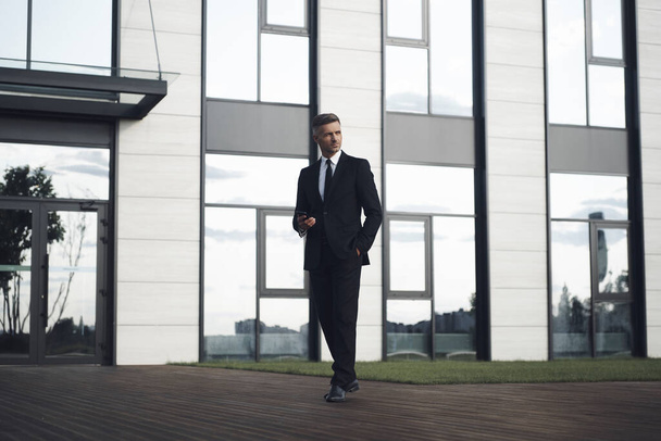 Confident mature businessman holding mobile phone while walking in front of office building - Fotoğraf, Görsel
