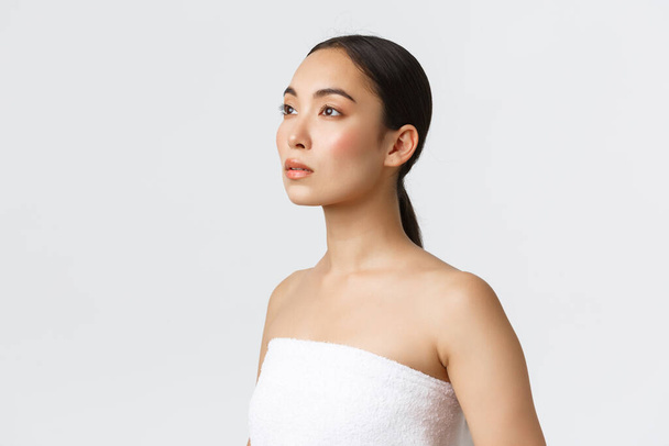 Kobiecy piękny azjatycki kobieta w białym ręczniku patrząc lewo marzycielski. Dziewczyna z czystą, idealną cerą, reklamą produktów do pielęgnacji skóry, kosmetyki łazienkowe, stojące białe tło - Zdjęcie, obraz