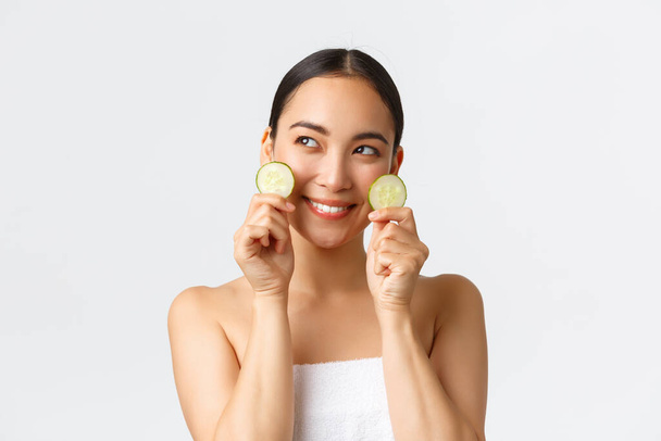 Beauty, personal care, spa salon and skincare concept. Coquettish beautiful asian girl in bath towel looking away dreamy with smile, showing cucumber pieces as promote face cream or mask - Фото, изображение