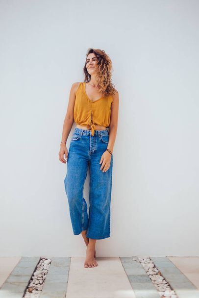 Barefoot female in yellow top and blue jeans standing on stone floor and leaning on white wall while looking away  - Fotó, kép