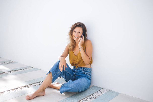 Barefooted smiling modern woman in summer orange top and blue jeans talking on mobile while sitting on marble floor leaning on white wall looking at camera - Fotografie, Obrázek