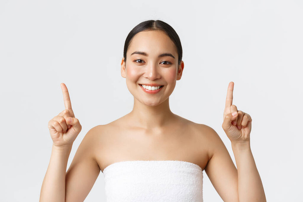 Schönheits-, Kosmetik- und Wellnesssalonkonzept. Glücklich zufrieden asiatische Frau im Handtuch lächelt zufrieden und zeigt mit den Fingern nach oben, empfehlen Massagetherapie, Hautpflegeprodukte, weißer Hintergrund - Foto, Bild