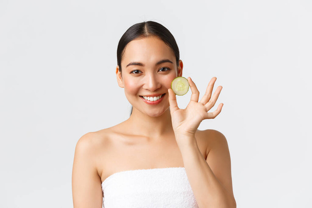 Beauty, personal care, spa salon and skincare concept. Beautiful asian female in bath towel holding cucumber piece over eye and smiling, promo of moisturizing face mask or cleanser, shower gel - Foto, immagini