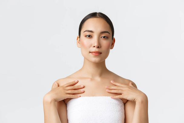 Tender beautiful asian woman with clean skin, perfect face, standing in towel and touching body, applying cream or cleanser, skincare products in bathroom, attend spa salon, white background - Foto, Imagem