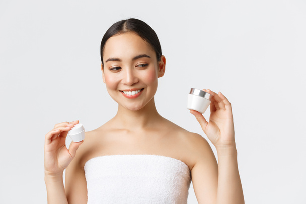 Schoonheid, persoonlijke verzorging, spa salon en huidverzorging concept. Close-up van mooie Aziatische vrouw in badhanddoek met twee crèmes, oog en gezicht voedende producten, glimlachen, huidbehandeling - Foto, afbeelding