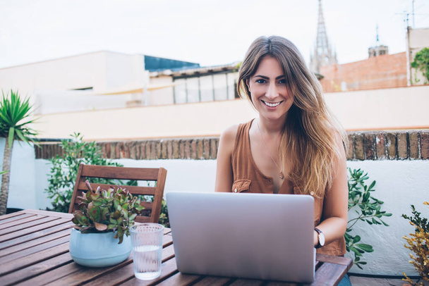 Happy young woman in brown shirt enjoying work outside sitting at wooden table with plant and surfing laptop while looking at camera - Foto, imagen