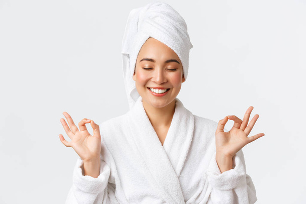 Personal care, women beauty, bath and shower concept. Close-up of relieved attractive asian woman attend spa beauty salon, wear bath towel and bathrobe, meditating, feeling nirvana, smiling - Photo, Image