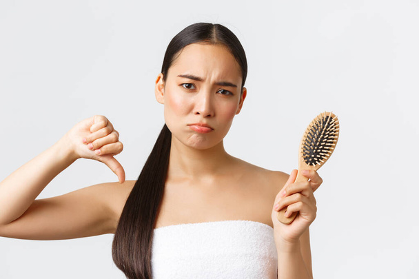 Beauty, treatment products, shampoo and hair care concept. Displeased sad pouting asian girl in bath towel frowning and showing thumb-down, complaining on hair loss, showing brush - Photo, Image