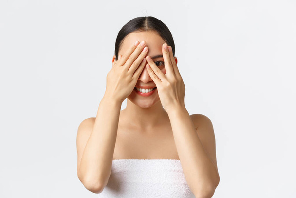 Beauty, cosmetology and spa salon concept. Silly beautiful asian girl in bath towel peeking through fingers as cover eyes with hands and smiling, interested what surprise she except, white background - Photo, Image