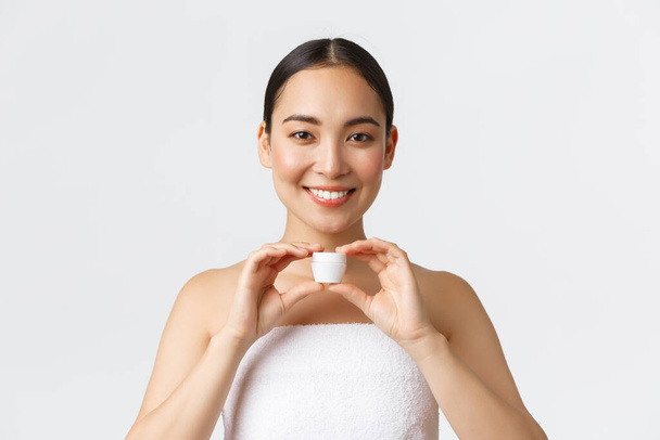 Bellezza, cura personale, salone termale e concetto di cura della pelle. Primo piano di bella donna asiatica sorridente in asciugamano mostrando crema per il viso, idratante, trattamento giornaliero del viso, sfondo bianco - Foto, immagini