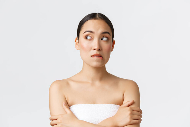 Beauty, cosmetology and spa salon concept. Doubtful and worried beautiful asian girl in towel biting lip and looking away indecisive, hesitating before wax depilation in beauty clinic - Photo, Image