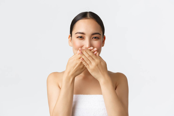 Beleza, cosmetologia e conceito de salão de spa. Bela mulher asiática em toalha escondendo lábios, cobrir a boca com as mãos e rir bobo, sorrindo com os olhos, de pé positivo sobre fundo branco
 - Foto, Imagem