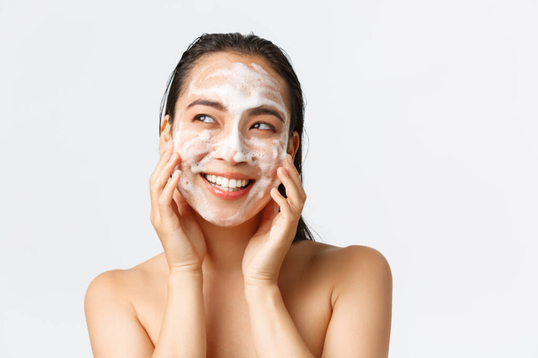 Skincare, women beauty, hygiene and personal care concept. Close-up of beautiful naked asian female smiling and looking left upbeat while clean skin with cleansing foam, daily care product - Fotoğraf, Görsel
