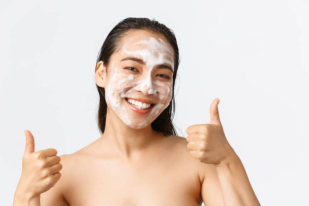 Pflege-, Schönheits-, Hygiene- und Pflegekonzept für Frauen. Nahaufnahme einer zufriedenen, lächelnden Asiatin, die nackt steht und Daumen nach oben zeigt, während sie Reinigungsschaum im Gesicht verwendet - Foto, Bild