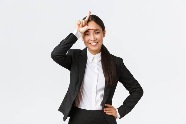 Успішна азіатська бізнес-леді в чорному костюмі знущається над своїми суперниками, показуючи знак невдахи як перемогу і святкування перемоги над конкурентами, стоячи задоволеними на білому тлі
 - Фото, зображення