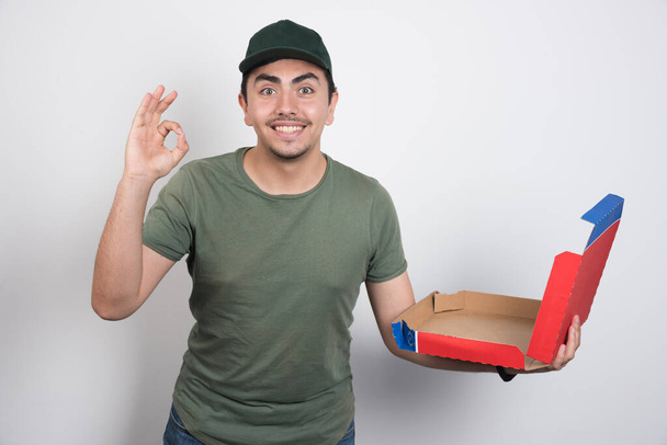 Deliveryman κάνει εντάξει σημάδι και κρατώντας άδειο κουτί σε λευκό φόντο. Υψηλής ποιότητας φωτογραφία - Φωτογραφία, εικόνα